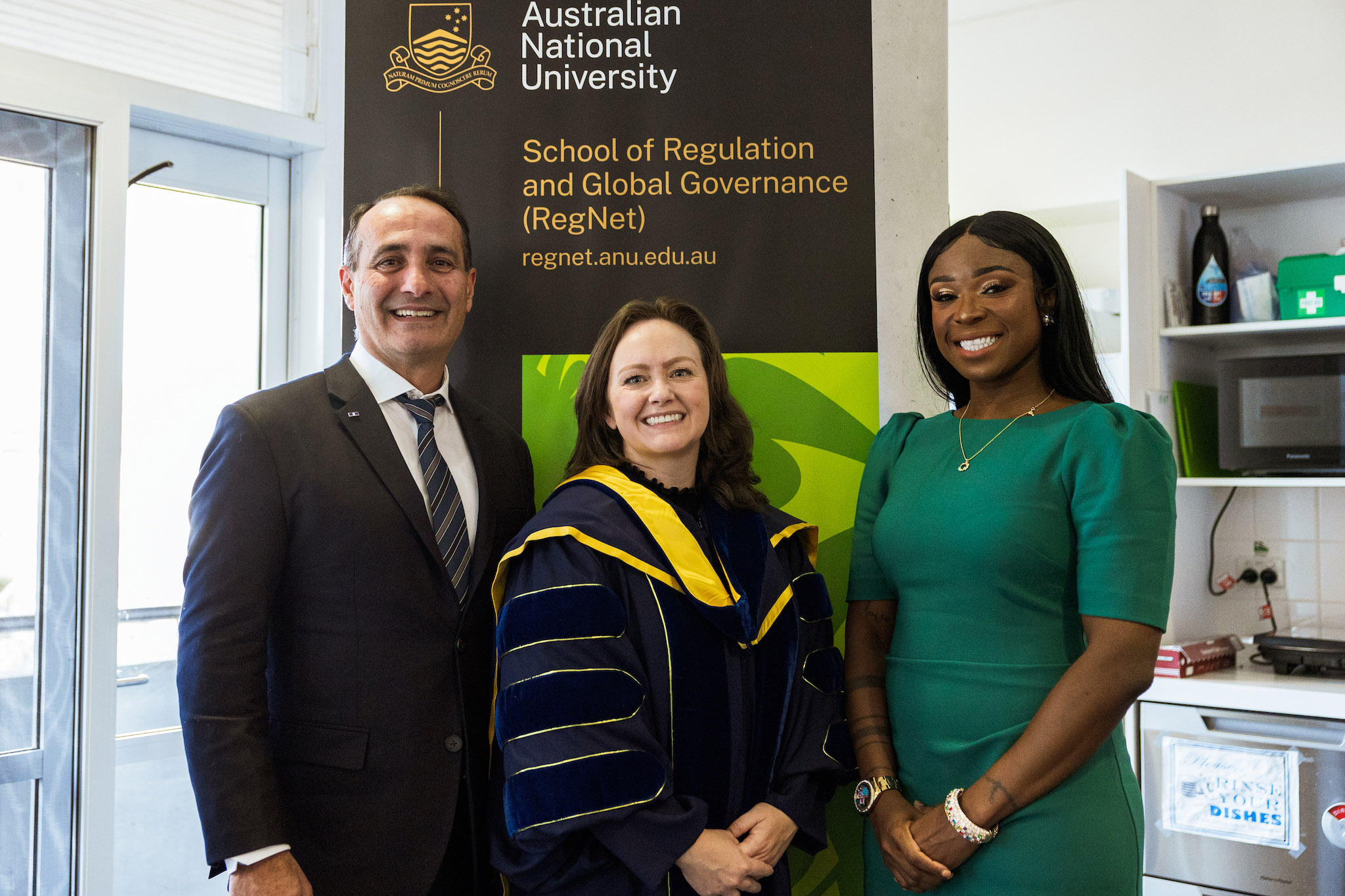 Graduate Steven Karras (left), Dean of RegNet School of Regulation and Global Governance Professor Kathryn Henne (middle) and graduate Precilla Lawrence Graduate Steven Karras (left), Dean of RegNet School of Regulation and Global Governance Professor Kathryn Henne (middle) and graduate Precilla Lawrence standing in front of a banner indoors